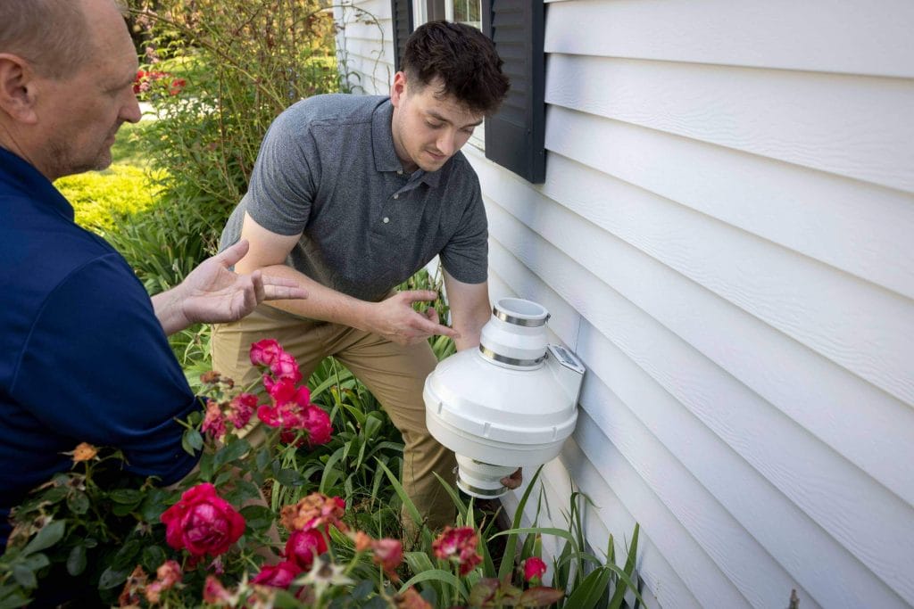 Two men putting in a radon mitigation system on someone's home exterior.