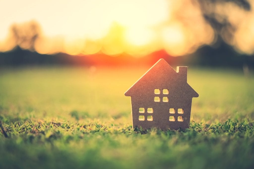 A wooden house cut out sitting on a grassy field with the sun shining in the background.