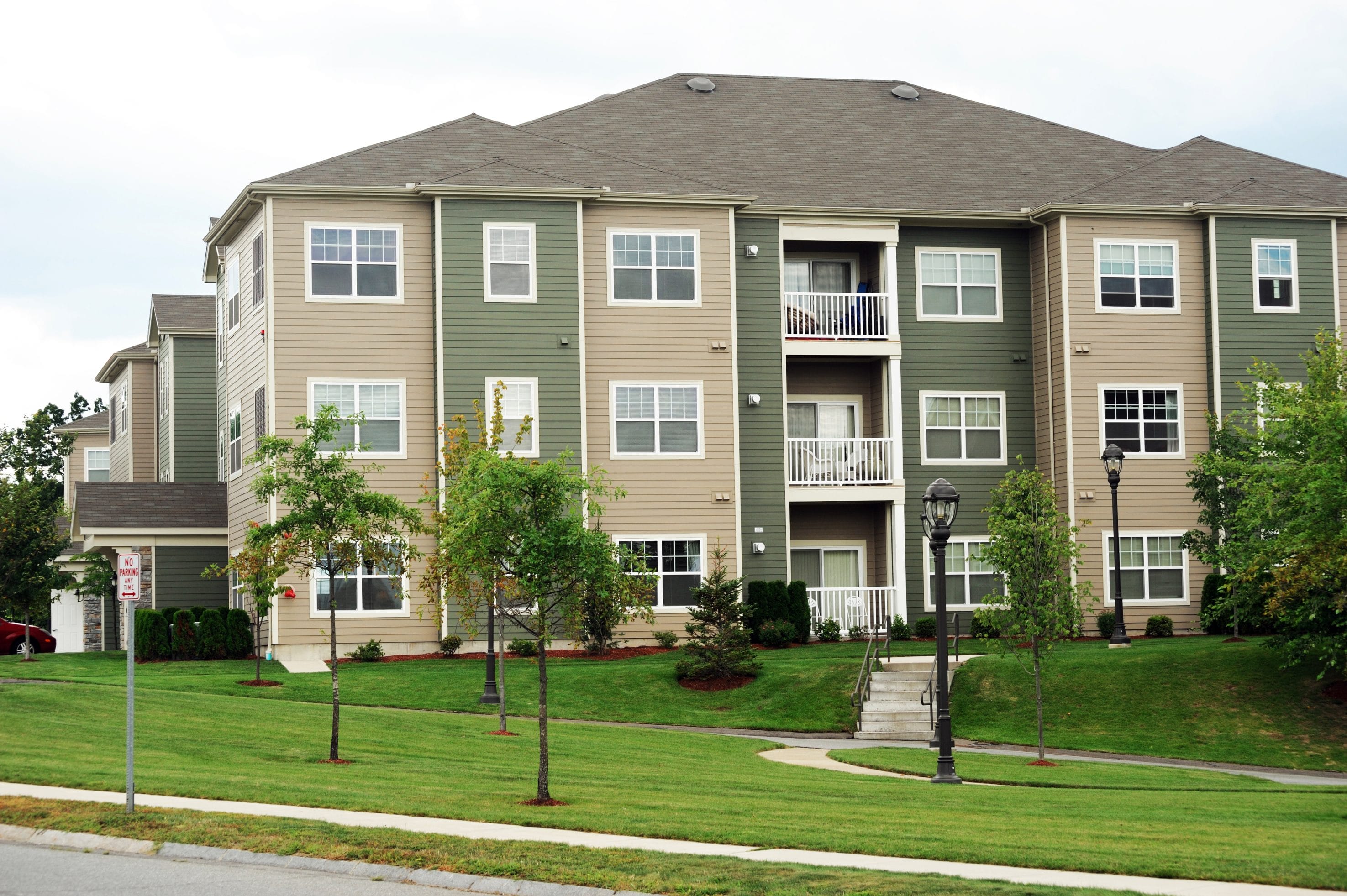 An exterior picture of a green and tan apartment complex with trees and bushes out front.